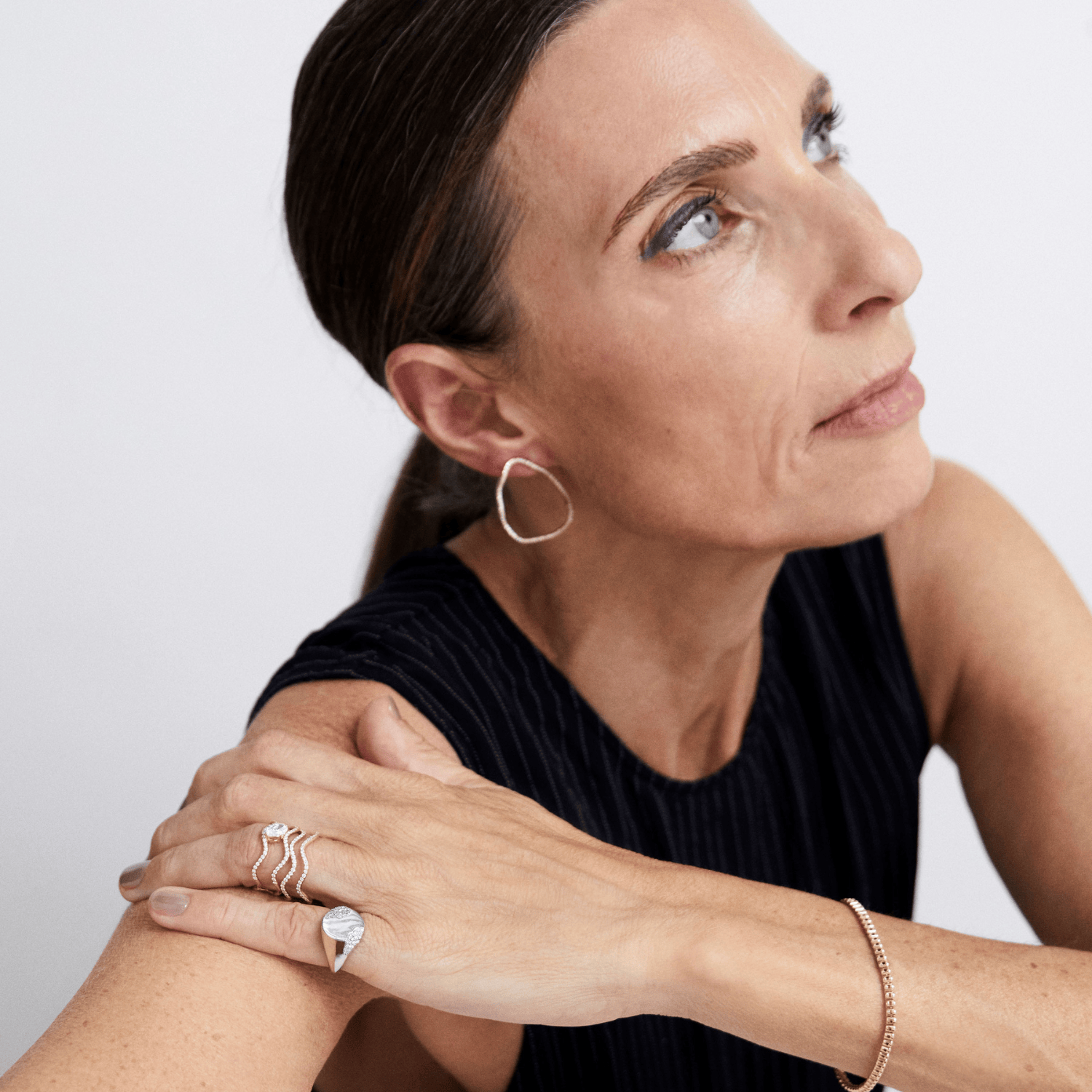 A close-up lifestyle shot of a woman wearing delicate gold jewelry, including a thin gold hoop earring, a gold chain bracelet, and multiple gold rings on one finger, against a plain light gray background.