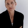 A close-up portrait of a woman with short blonde hair wearing delicate gold dangle earrings, a black blazer, and a white top, set against a plain light gray background.