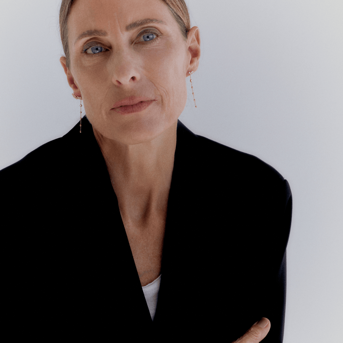 A close-up portrait of a woman with short blonde hair wearing delicate gold dangle earrings, a black blazer, and a white top, set against a plain light gray background.