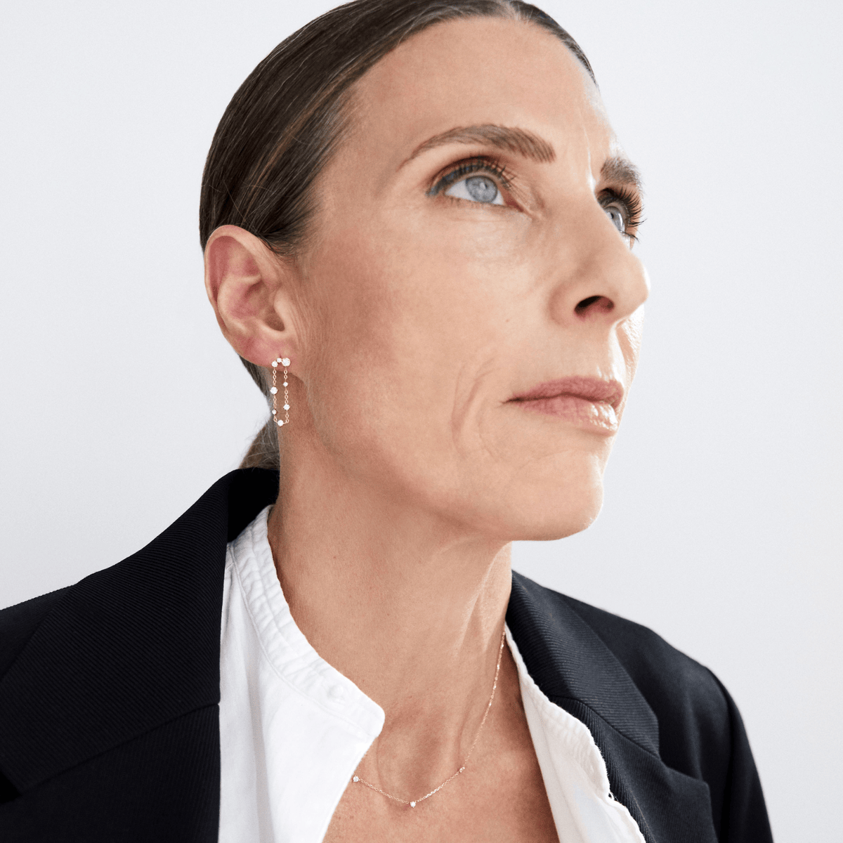 A close-up portrait of a woman wearing delicate gold hoop earrings, a fine gold necklace, and a black blazer over a white collared shirt, set against a plain light gray background.