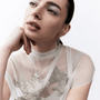 A close-up portrait of a woman wearing a sheer white mock-neck top with silver sequined leaf patterns and a dangling silver hoop earring, set against a plain white background.