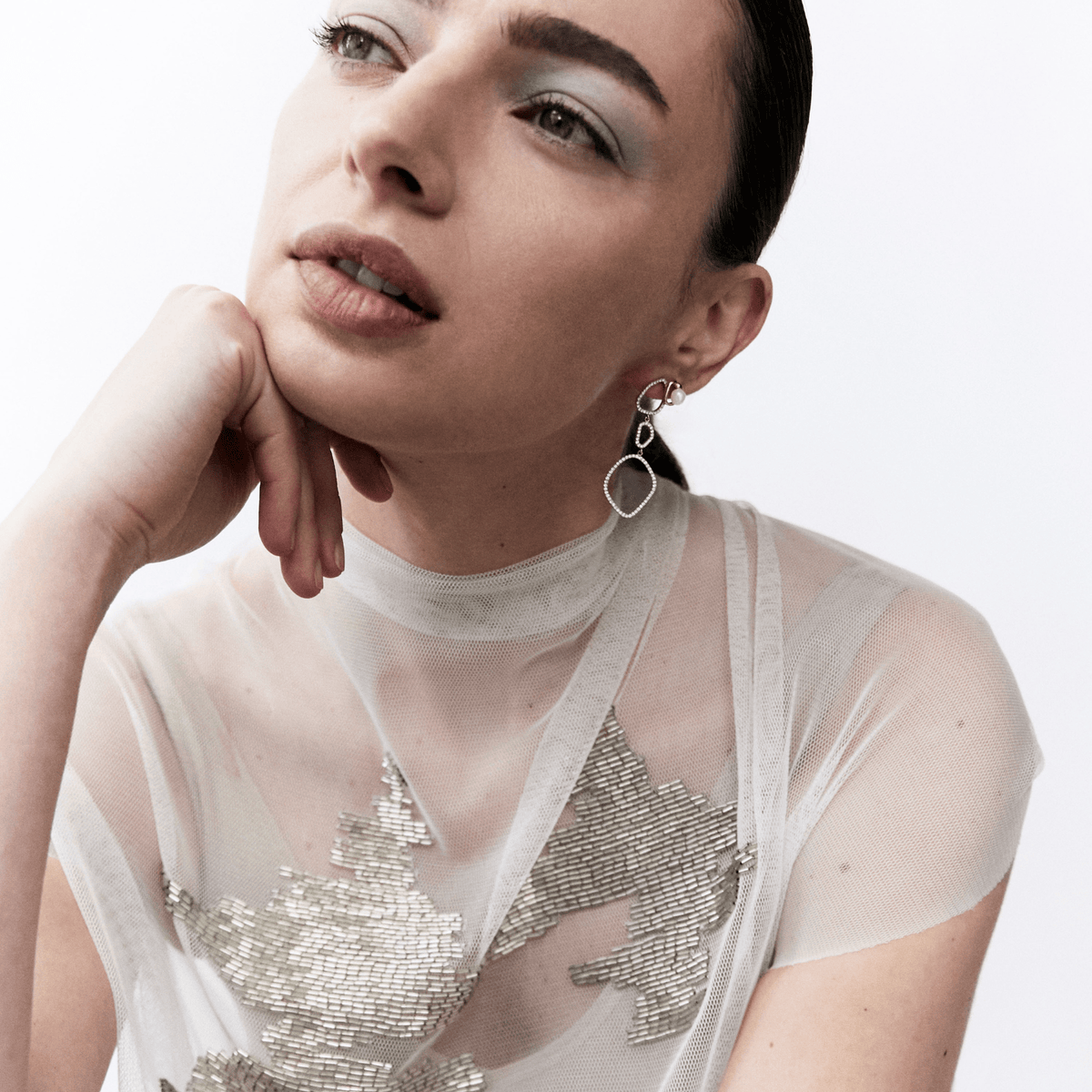 A close-up portrait of a woman wearing a sheer white mock-neck top with silver sequined leaf patterns and a dangling silver hoop earring, set against a plain white background.