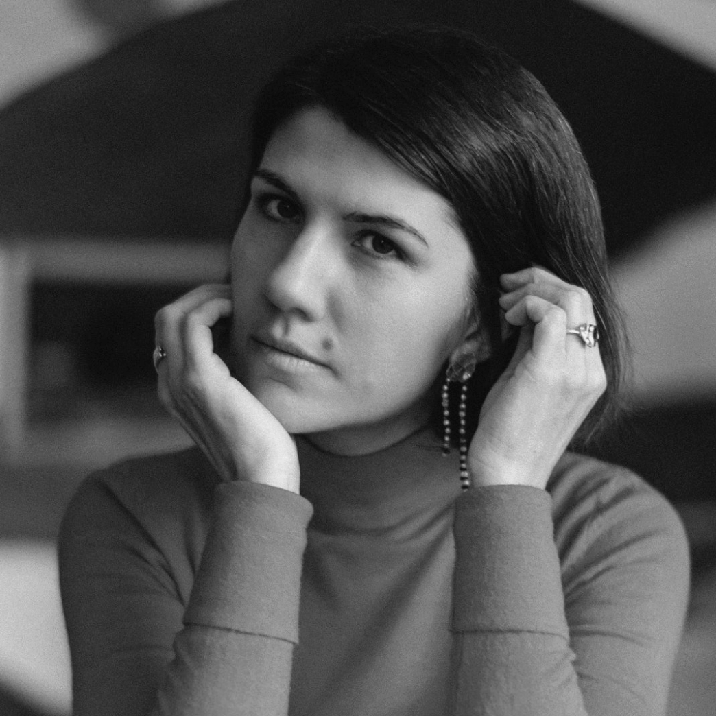 A black and white portrait of a woman with dark hair, wearing a turtleneck sweater and dangling earrings, resting her chin on her hands.