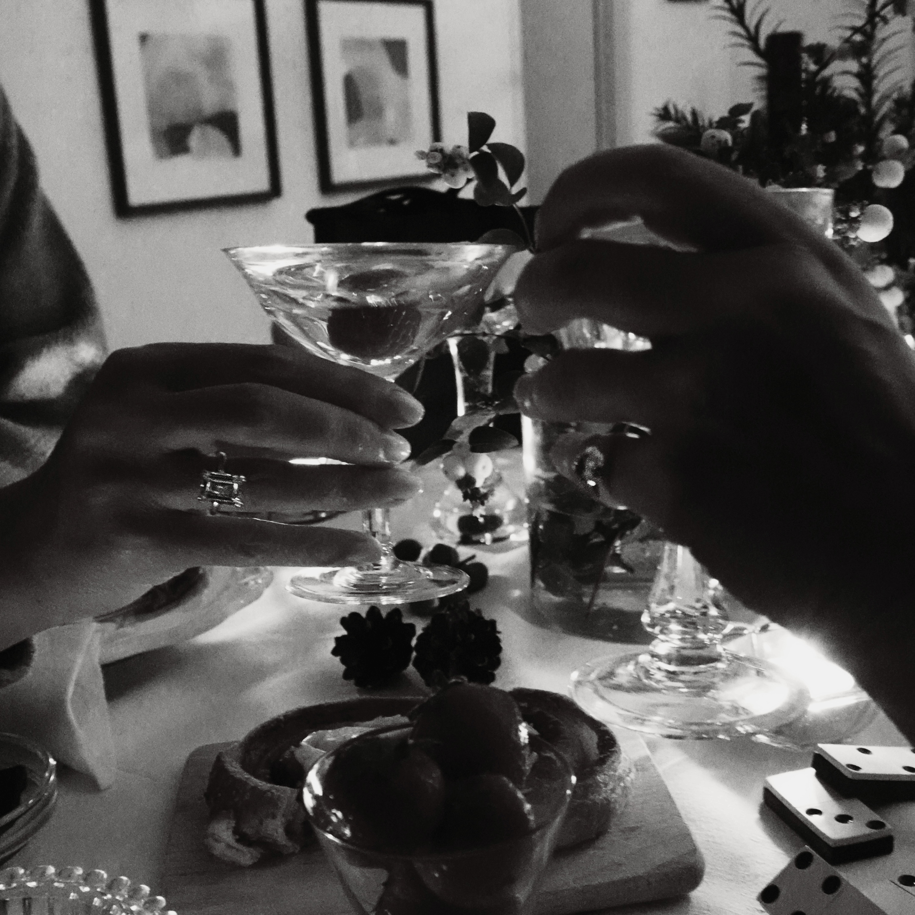 A black and white lifestyle photograph of two hands clinking martini glasses over a dining table with olives, framed pictures on the wall, and dice.